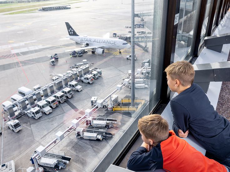 Zwei Kinder stehen an einer großen Fensterfront im Terminal des Flughafens Hannover und schauen auf das Vorfeld mit Flugzeug und Abfertigungsfahrzeugen.