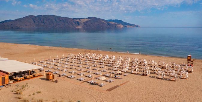 Weitläufiger Sandstrand am Hotel Georgioupolis Suites auf Kreta mit zahlreichen Sonnenliegen und Sonnenschirmen, Blick auf das ruhige Meer und eine bergige Küstenlandschaft im Hintergrund bei klarem Himmel