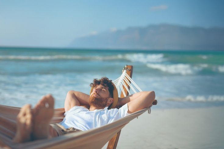 Person liegt entspannt in einer Hängematte am Strand, trägt weißes T-Shirt, im Hintergrund Meer und Berge.