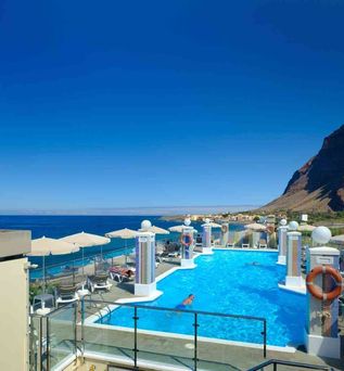 Außenpool mit mehreren weißen Sonnenschirmen, Liegestühlen und einer Person im Wasser, dahinter Meer und Küstenlinie unter blauem Himmel.