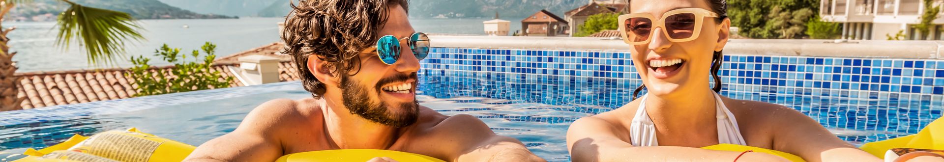 Lachendes Paar entspannt im Pool auf einer gelben Luftmatratze, vor mediterraner Kulisse mit Meerblick und sonniger Urlaubsatmosphäre