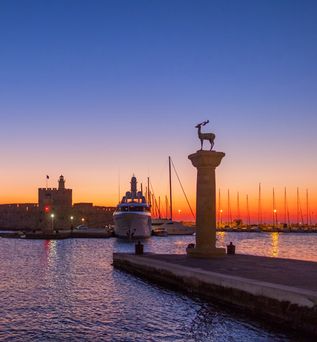 Blick auf den Mandraki-Hafen in Rhodos bei Sonnenuntergang mit zwei Säulen, von denen eine eine Hirschstatue trägt, und einem großen Schiff am Kai.