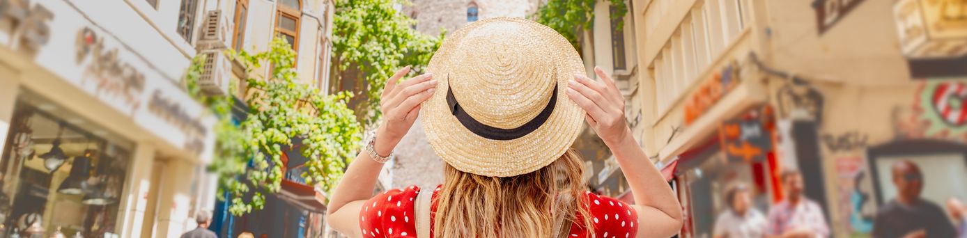 Frau mit Strohhut und langen Haaren steht in einer belebten Einkaufsstraße mit Geschäften und Cafés