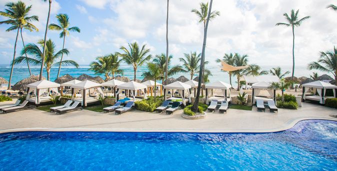 Pool mit Liegen und Sonnenschirmen vor Palmen und Meer unter blauem Himmel, direkt davor das Meer.