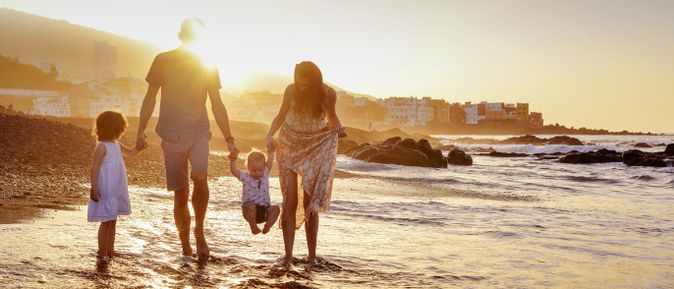 Familie spaziert bei Sonnenuntergang am Strand entlang, die Eltern halten ihre Kinder an der Hand