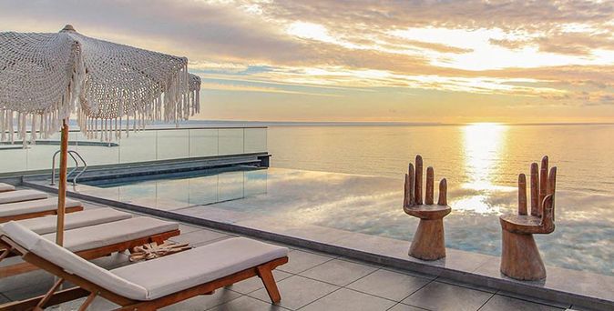 Holzliegen unter weißen Sonnenschirmen am Infinity-Pool mit Blick auf Sonnenuntergang über dem Meer