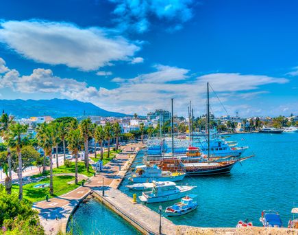 Hafen von Kos mit mehreren Segel- und Motorbooten an der Mole, Palmen und Gebäuden im Hintergrund unter blauem Himmel mit Wolken