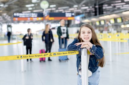 Kind lehnt sich hinter einem gelben Absperrband mit der Aufschrift „Köln Bonn Airport“ im Check-in-Bereich des Flughafens Köln/Bonn an einen Absperrpfosten