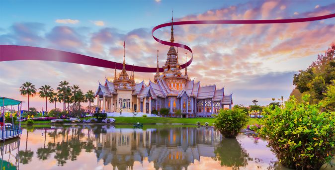 Thailändischer Tempel mit spitzen Dächern und goldenen Verzierungen, umgeben von Wasser und Palmen, bei Sonnenuntergang.