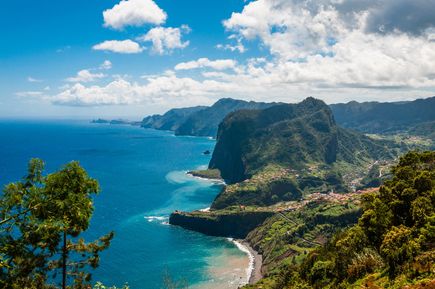 Panoramablick auf die Küste von Faial auf Madeira mit grünen Steilhängen, dem Atlantik, einer geschwungenen Bucht und kleinen Ortschaften entlang der Küste unter einem wolkenreichen Himmel