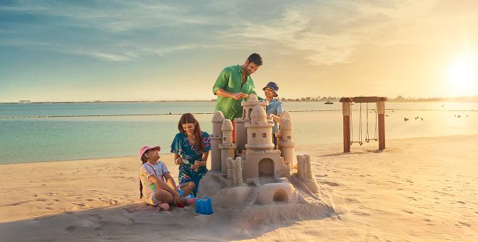 Familie baut große Sandburg am Strand von Ras Al Khaimah bei Sonnenuntergang, im Hintergrund Meer und Schaukel.