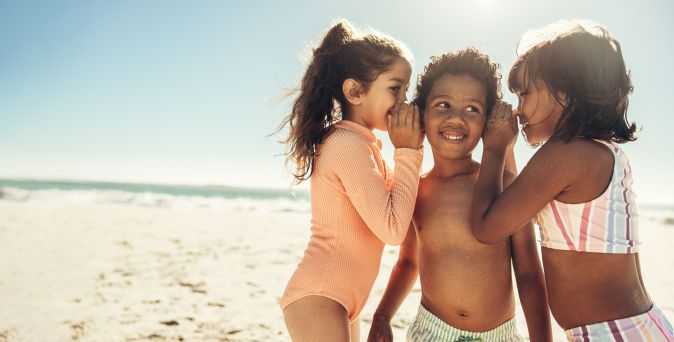 Drei Kinder am Strand, zwei flüstern einem lachenden Kind etwas ins Ohr