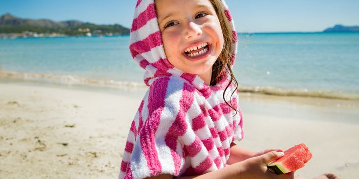 Fröhliches Kind im gestreiften Handtuch-Poncho isst ein Stück Wassermelone am Strand