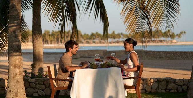 Zwei Personen sitzen an einem mit weißem Tuch bedeckten Tisch unter Palmen am Strand, mit Getränken und Speisen auf dem Tisch im Juweira Boutique Hotel in Salalah, Oman.