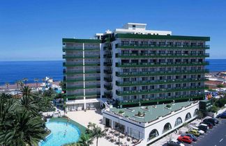 Großer runder Hotelpool mit Liegestühlen und Palmen, angrenzendes Hotelgebäude links, Hügel mit Häusern und Meer im Hintergrund unter blauem Himmel.