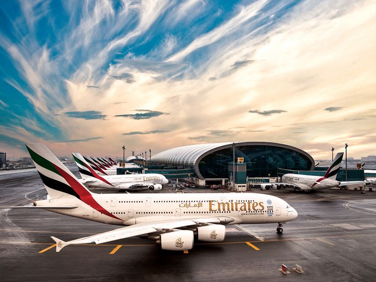 Mehrere Emirates Flugzeuge stehen vor dem modernen Terminalgebäude des Dubai International Airport, während ein Emirates Airbus A380 im Vordergrund auf dem Rollfeld steht