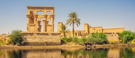 Philae-Tempel mit Säulen und Ruinen am Ufer eines Flusses, Palmen und klarer Himmel im Hintergrund