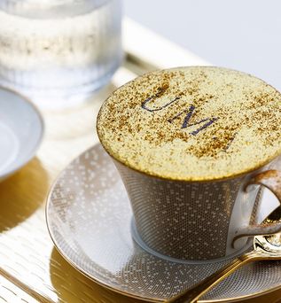 Kaffeetasse mit Milchschaum und Blattgold, auf dem 'UMI' steht, auf Untertasse mit Muster, daneben goldfarbener Löffel und kleines Gebäck auf weißem Teller.