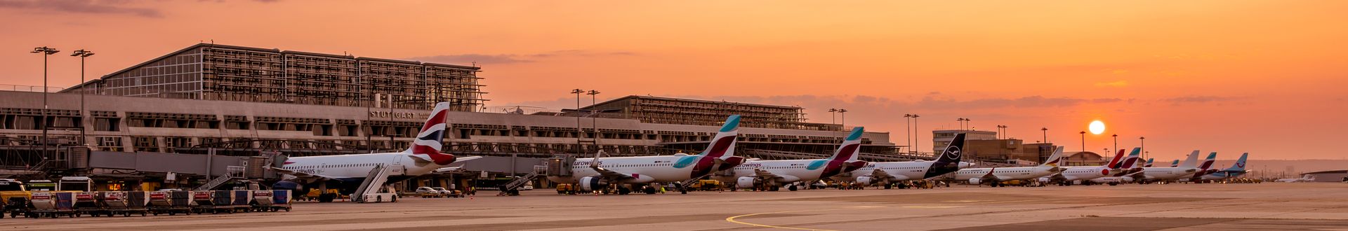 Am Flughafen Stuttgart stehen mehrere Passagierflugzeuge verschiedener Airlines vor dem Terminal, während die Sonne bei Sonnenuntergang über dem Vorfeld zu sehen ist