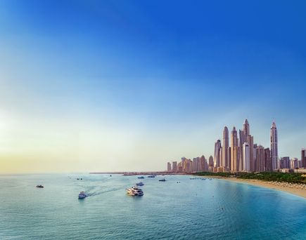 Blick auf die Skyline der Dubai Marina mit ihren modernen Hochhäusern, einer geschwungenen Sandbucht und mehreren Booten, die über das türkisfarbene Wasser fahren, unter klarem blauem Himmel