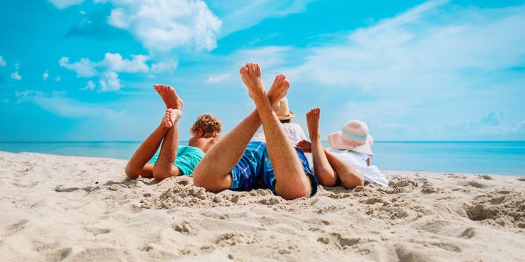 Erwachsene Person und zwei Kinder liegen entspannt auf dem Bauch im Sand am Strand mit Blick aufs Meer