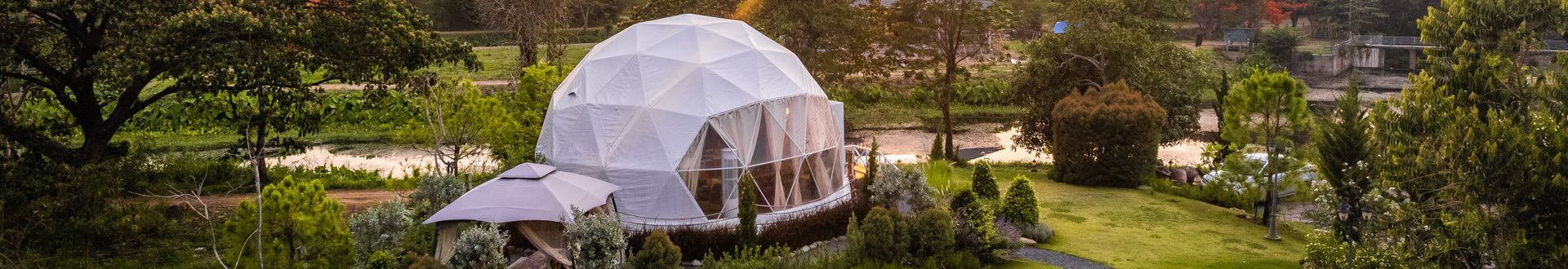 Glamping-Dome in grüner Landschaft mit geometrischer Kuppel, umgeben von Bäumen, Rasenflächen und einem Fluss im Hintergrund