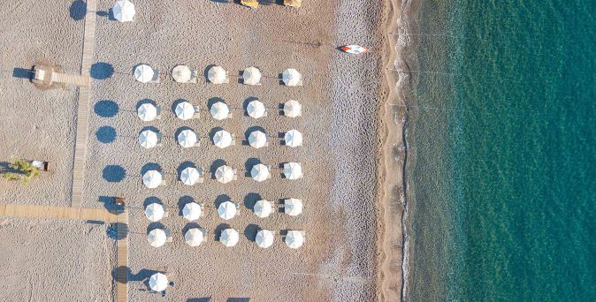 Luftaufnahme eines Strandes mit mehreren Reihen von Sonnenschirmen und Liegen neben klarem Wasser