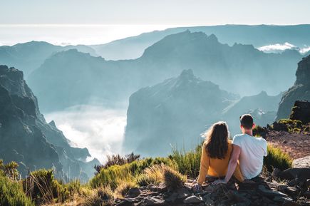 Personen sitzen auf einem Aussichtspunkt am Pico do Arieiro auf Madeira und blicken über die von Wolken umgebenen Berggipfel der Insel