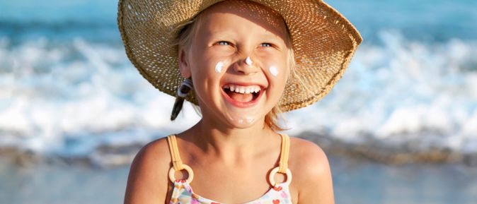 Fröhliches Kind mit Strohhut am Strand, lachend in die Sonne blickend, mit Sonnencreme auf Wangen und Nase