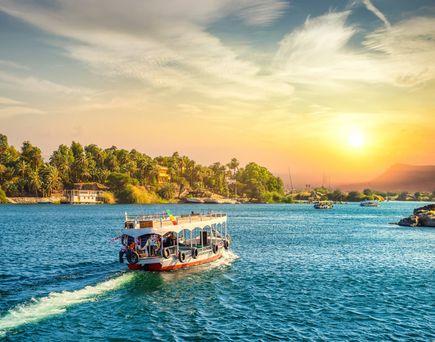 Boot mit offenem Deck auf dem Nil bei Sonnenuntergang mit Palmen und Gebäuden am Ufer