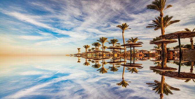 Spiegelglatte Wasseroberfläche reflektiert Palmen und Strohdach-Sonnenschirme unter blauem Himmel mit leichten Schleierwolken in Hurghada, Ägypten.