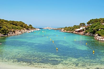 Ansicht der Bucht Cala Santandria auf Menorca mit seichtem, türkisblauem Wasser, gelben Bojen und Booten vor grüner Küste