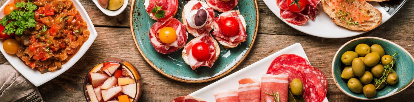 Verschiedene mediterrane Vorspeisen auf Tellern, darunter Oliven, Tomaten mit Mozzarella, Aufschnitt und Brot.