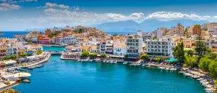 Blick auf den Hafen von Agios Nikolaos auf Kreta mit bunten Gebäuden, Booten und Bergen im Hintergrund.