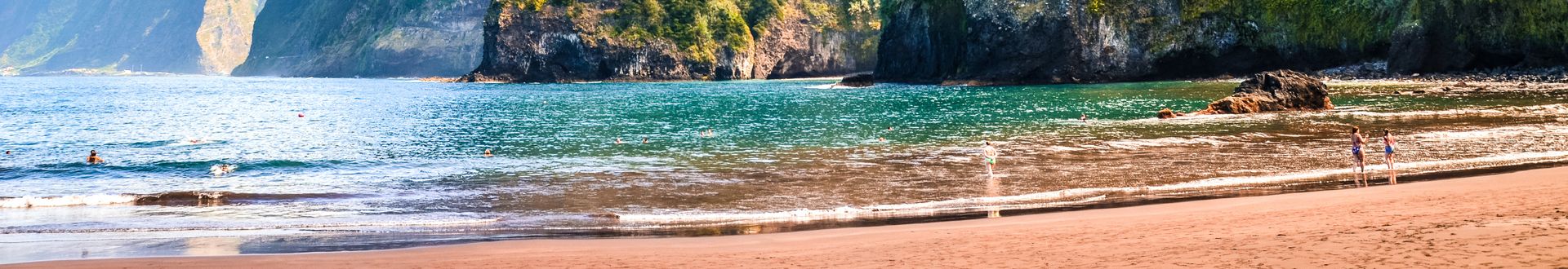 Panoramablick auf den Sandstrand Praia do Porto do Seixal auf Madeira mit Badegästen am Ufer, türkisgrünem Atlantik, felsiger Küste und steilen, dicht bewachsenen Berghängen im Hintergrund