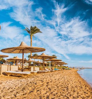 Strand am Roten Meer in Ägypten mit strohbedeckten Sonnenschirmen, Liegen und ruhigem, flach abfallendem Wasser unter blauem Himmel