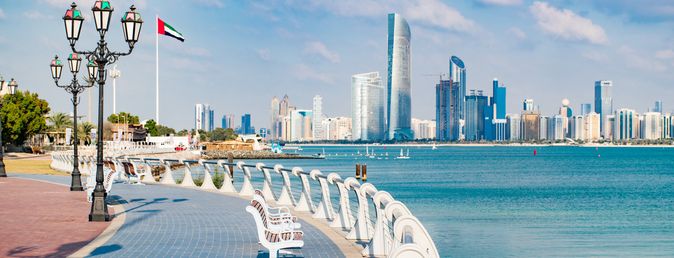 Skyline mit modernen Wolkenkratzern, Sitzbänken entlang der Uferpromenade und einer Flagge der Vereinigten Arabischen Emirate an einer Laterne