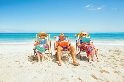 Drei Personen sitzen in bunten Liegestühlen am sonnigen Sandstrand und lesen Bücher, mit Blick auf das türkisfarbene Meer und den klaren blauen Himmel