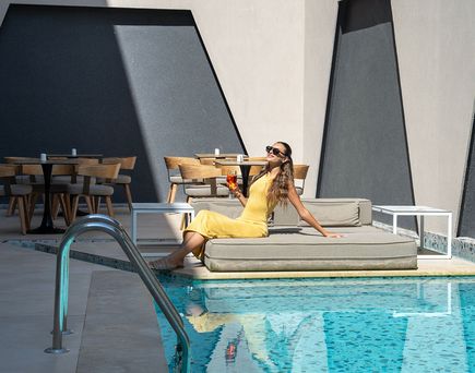 Frau in gelbem Kleid sitzt auf grauem Polster neben Pool im Hotel Zorbas Lifestyle, umgeben von modernem, geometrischem Design und Holztischen.