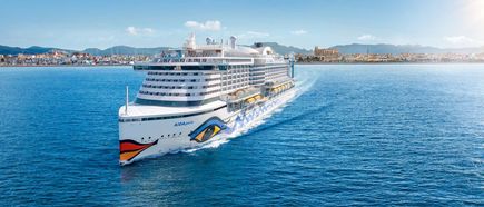 Das Kreuzfahrtschiff AIDAperla fährt bei sonnigem Wetter über das tiefblaue Meer, mit einer Küstenstadt und Gebirgslandschaft im Hintergrund