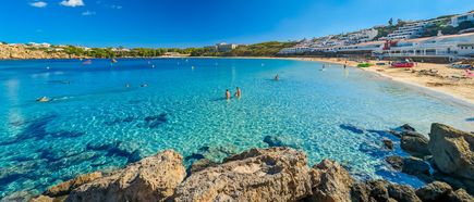 Halbrunde Badebucht Arenal d’en Castell auf Menorca mit klarem Wasser, Sandstrand und Ferienhäusern am Hang