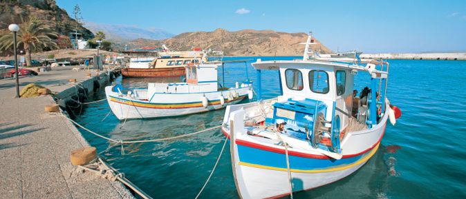 Fischerboote mit weißen Rümpfen und bunten Streifen liegen an einem Hafenpier vor einer felsigen Küstenlandschaft unter blauem Himmel.