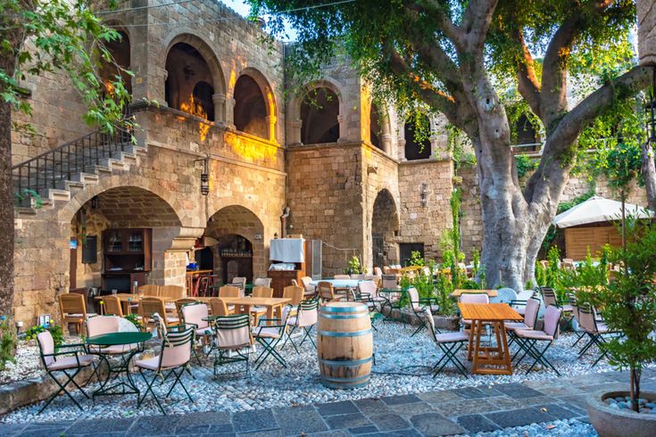 Innenhof eines Restaurants mit Tischen, Stühlen, einem Weinfass und großen Bäumen vor einem historischen Gebäude mit Arkaden