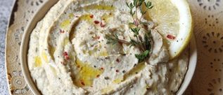 Schale mit cremigem Auberginen-Dip, garniert mit Olivenöl, roten Pfefferkörnern, Thymianzweigen und einer Zitronenscheibe auf einem gemusterten Teller.