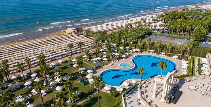 Luftaufnahme eines Hotels mit großem Pool, Liegen, Sonnenschirmen, Palmen und Strand mit Wellen im Hintergrund