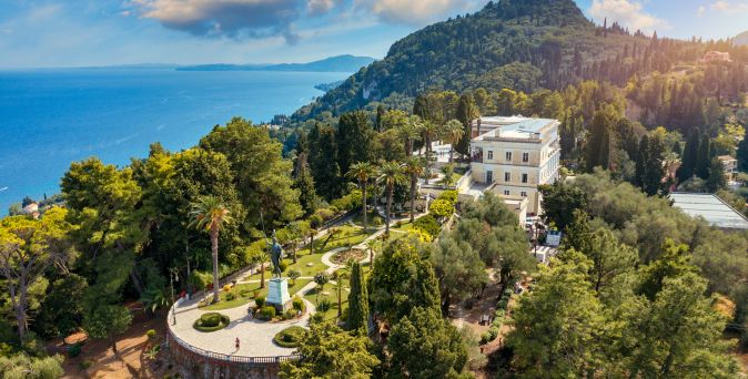 Luftaufnahme des Achilleion-Palasts mit umliegendem Garten, Palmen und Blick auf das Meer bei klarem Himmel.
