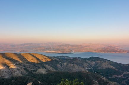 Berglandschaft mit Hügeln im Vordergrund und weiter entfernten Bergen am Meer unter klarem Himmel