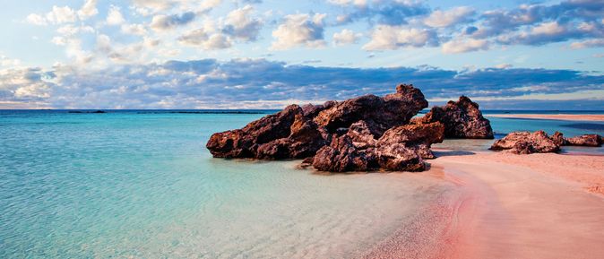 Rosa Sandstrand mit sanften Wellen und großen Felsen im Wasser, unter einem Himmel mit Wolken. Aufnahme von Kreta.