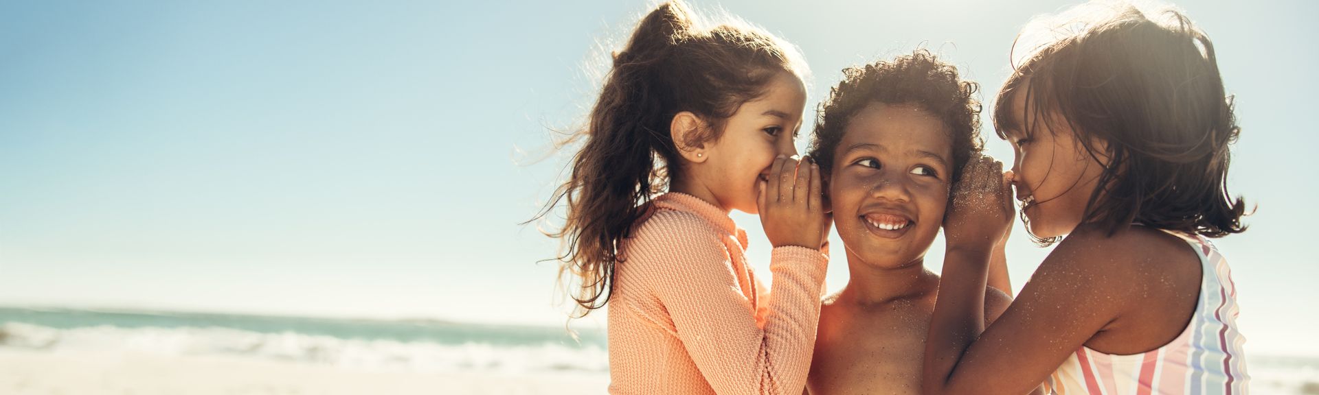 Drei Kinder am Strand, zwei flüstern einem lachenden Kind etwas ins Ohr