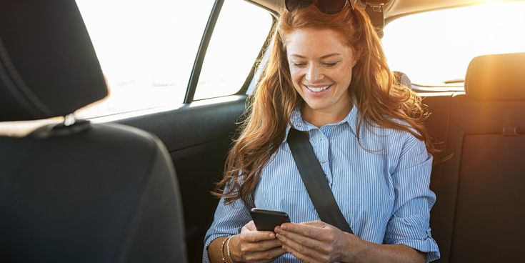 Frau sitzt angeschnallt auf dem Rücksitz eines Autos und lächelt auf ihr Smartphone, warmes Sonnenlicht fällt durch das Fenster